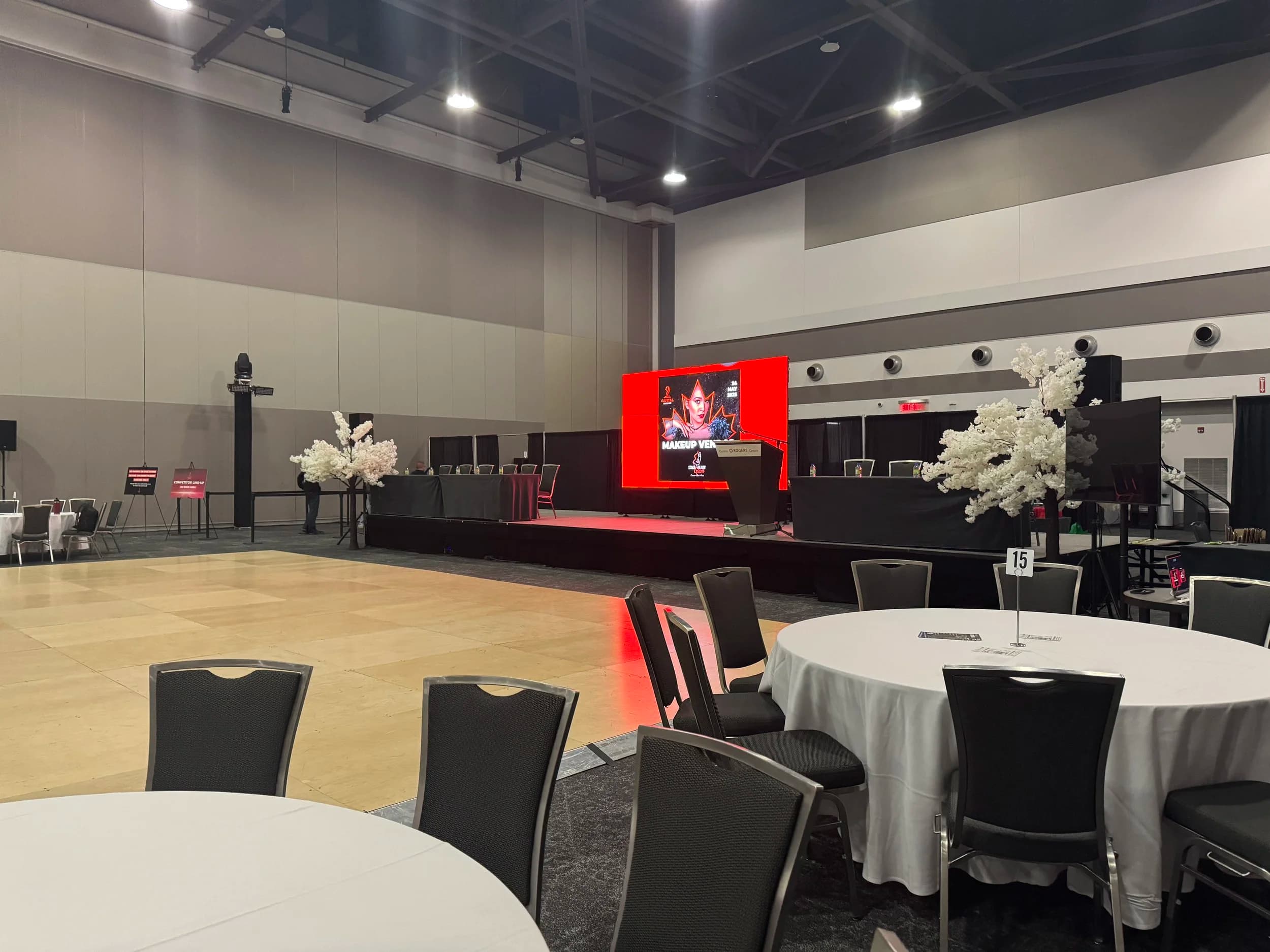 Wide view of a Rogers Centre Ottawa ballroom set for a competition expo — branded LED wall on stage, white blossom trees, banquet seating ready for doors